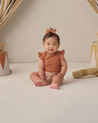 Baby sitting on a light-colored floor wearing a rust-colored outfit with ruffled sleeves.