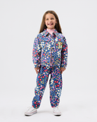 A young girl stands against a light gray studio background wearing a matching long-sleeve button-front jumpsuit with an all-over colorful floral print on a dark blue base, along with white sneakers.