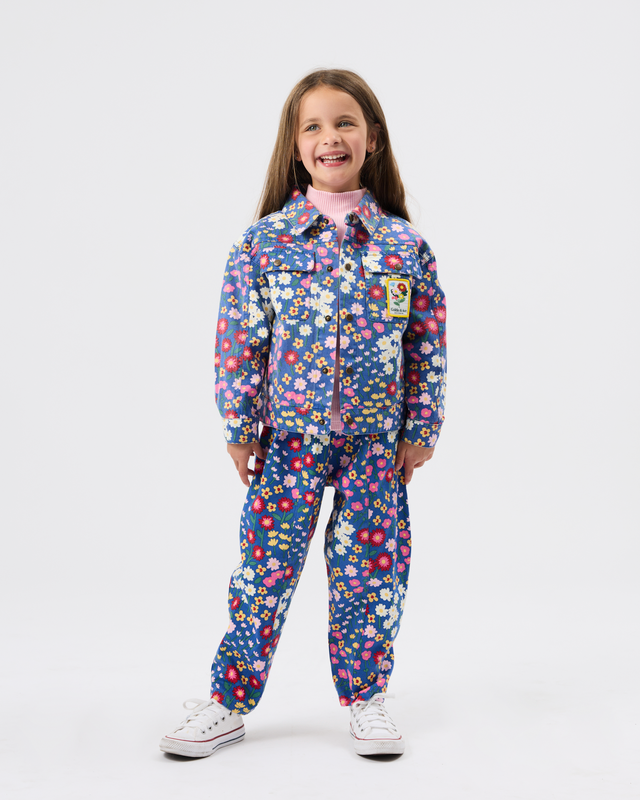 A young girl stands against a light gray studio background wearing a matching long-sleeve button-front jumpsuit with an all-over colorful floral print on a dark blue base, along with white sneakers.
