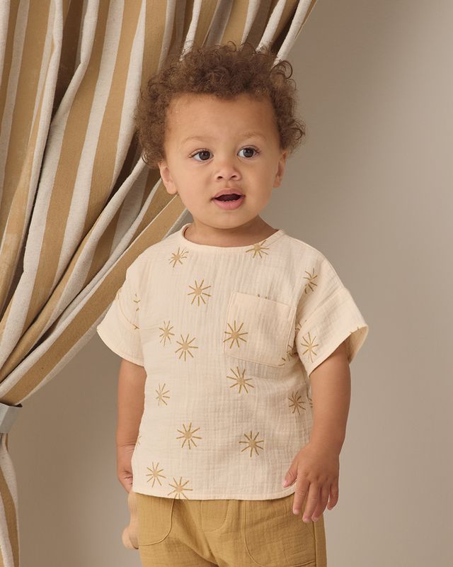 Child wearing a beige shirt with gold star patterns in front of a curtain.