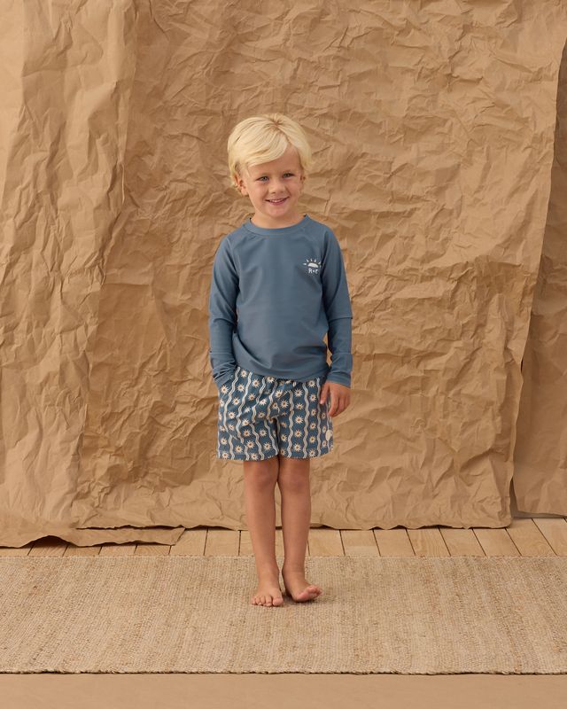 Child wearing a blue shirt and patterned shorts standing against a textured brown backdrop