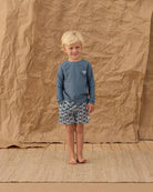 Child wearing a blue shirt and patterned shorts standing against a textured brown backdrop
