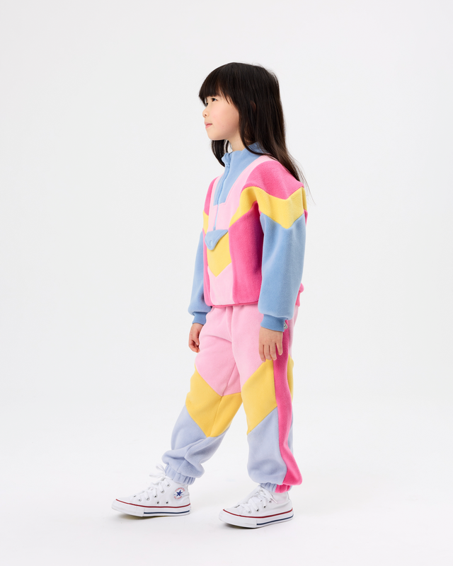 A child model wearing a pastel color-blocked tracksuit with pink, light blue, and yellow geometric panels, paired with white sneakers, photographed against a seamless white studio background.