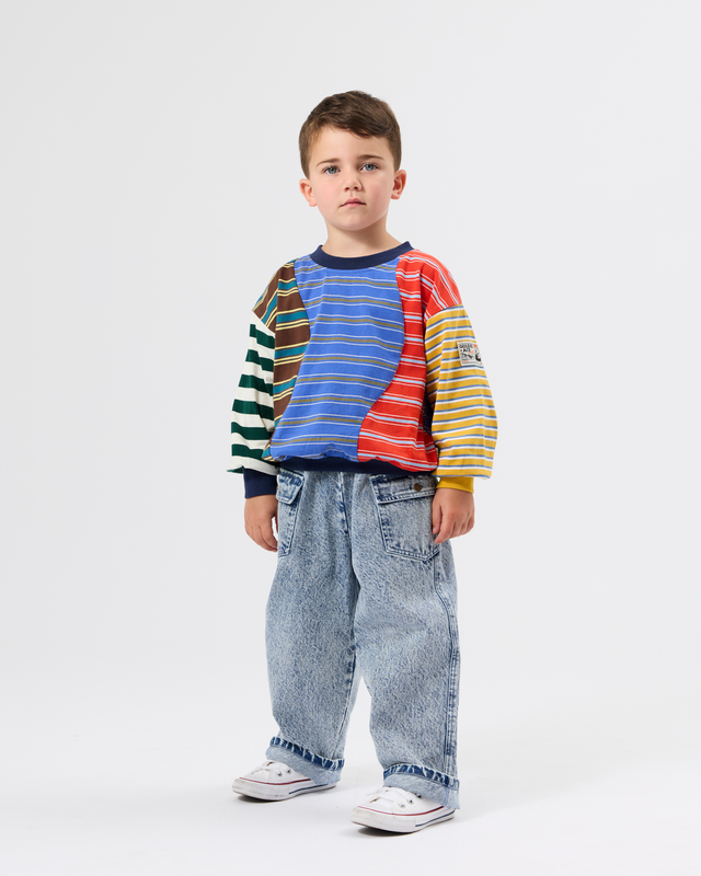 A young boy stands against a light gray studio background wearing a colorful striped long-sleeve shirt with blue jeans and white sneakers.