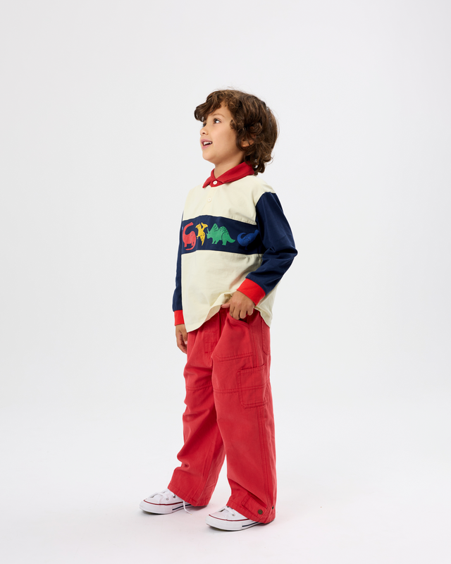 A young boy wearing a color-blocked long-sleeve sweater with dinosaur graphics, red cargo pants, and white sneakers, standing in profile against a plain white studio background.