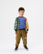 A young boy modeling a colorful long-sleeve shirt with striped panels on the sleeves and chest, paired with brown pants and navy sneakers, against a plain white studio background.