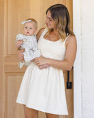 Woman in a white dress holding a baby in front of a wooden door.