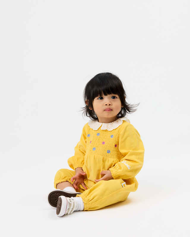 A toddler girl with short black hair sits on a white studio floor wearing a yellow smocked romper with a white ruffled collar and small colorful embroidered dots.