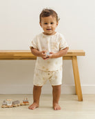 Child wearing a white outfit with gold patterns standing in front of a wooden table.