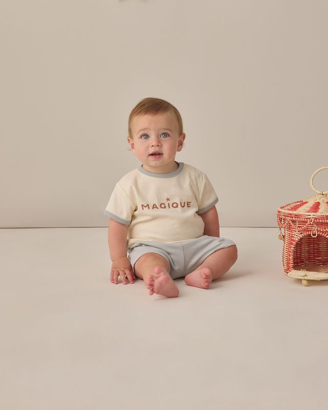 Baby sitting on a plain background wearing a 'Magique' shirt and gray shorts.