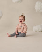 Child wearing a pink sweatshirt with a cherry design, sitting on a beige floor with white cloud decorations.