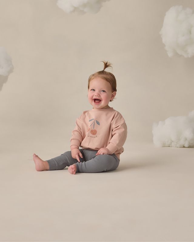Child wearing a pink sweatshirt with a cherry design, sitting on a beige floor with white cloud decorations.