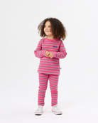 A young girl with curly brown hair stands against a white studio background wearing a matching pink and light blue striped long-sleeve top and pants set, paired with white sneakers.