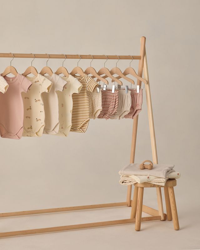 Row of baby rompers on a wooden rack with a stool underneath against a beige background