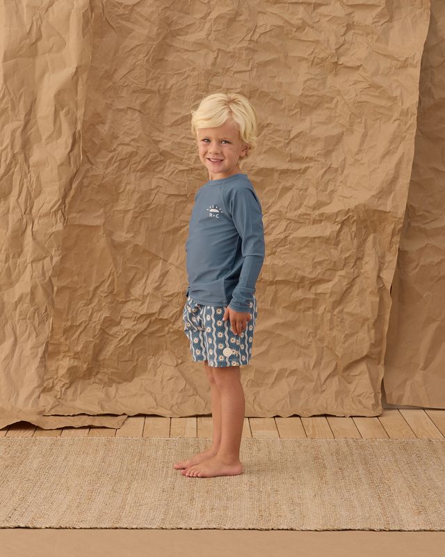 Child wearing a blue shirt and patterned shorts standing against a textured brown backdrop.