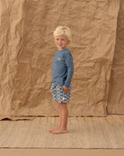 Child wearing a blue shirt and patterned shorts standing against a textured brown backdrop.