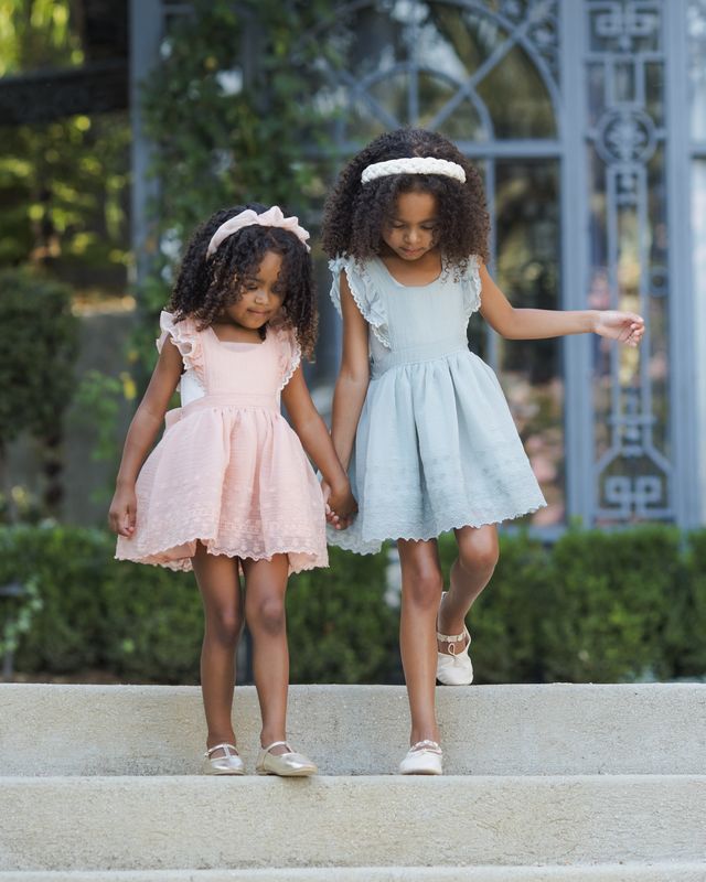 Two young girls in dresses standing on a stone ledge with greenery in the background