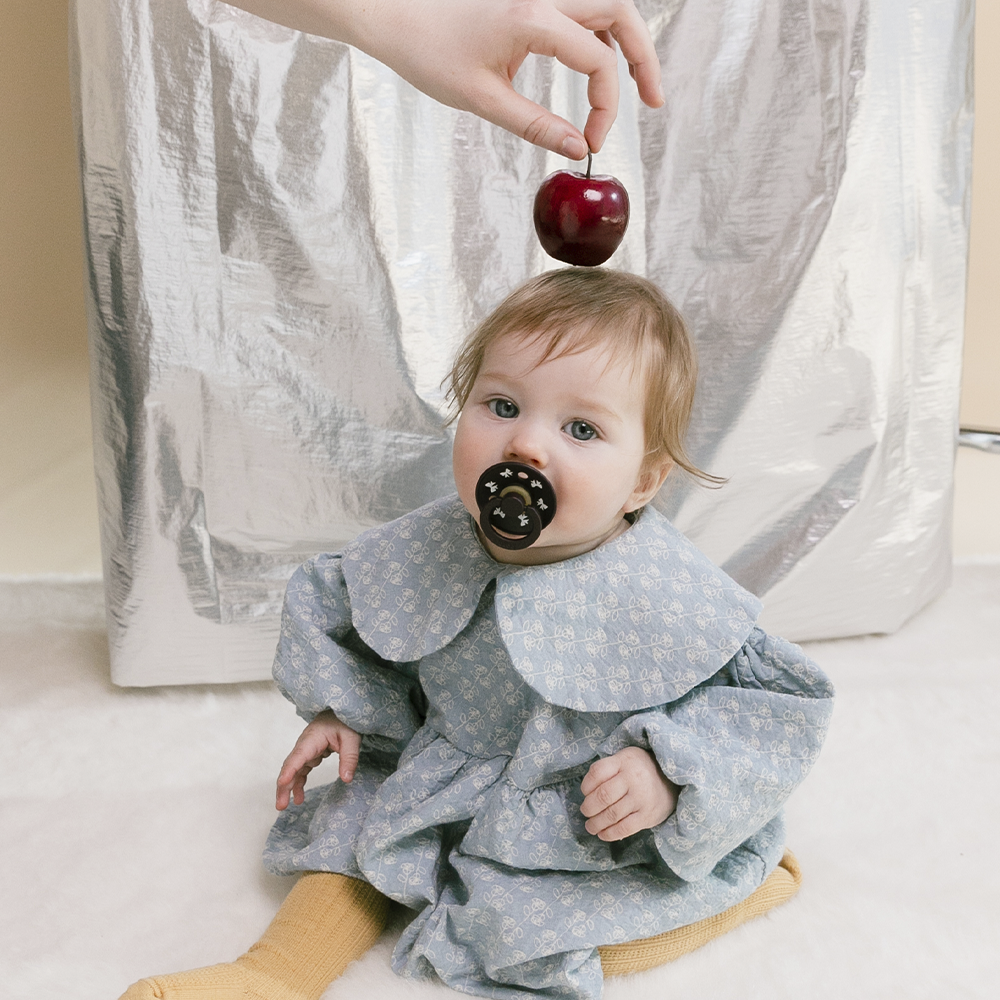Baby in a light blue outfit with a dark pacifier, holding an apple, against a neutral background.
