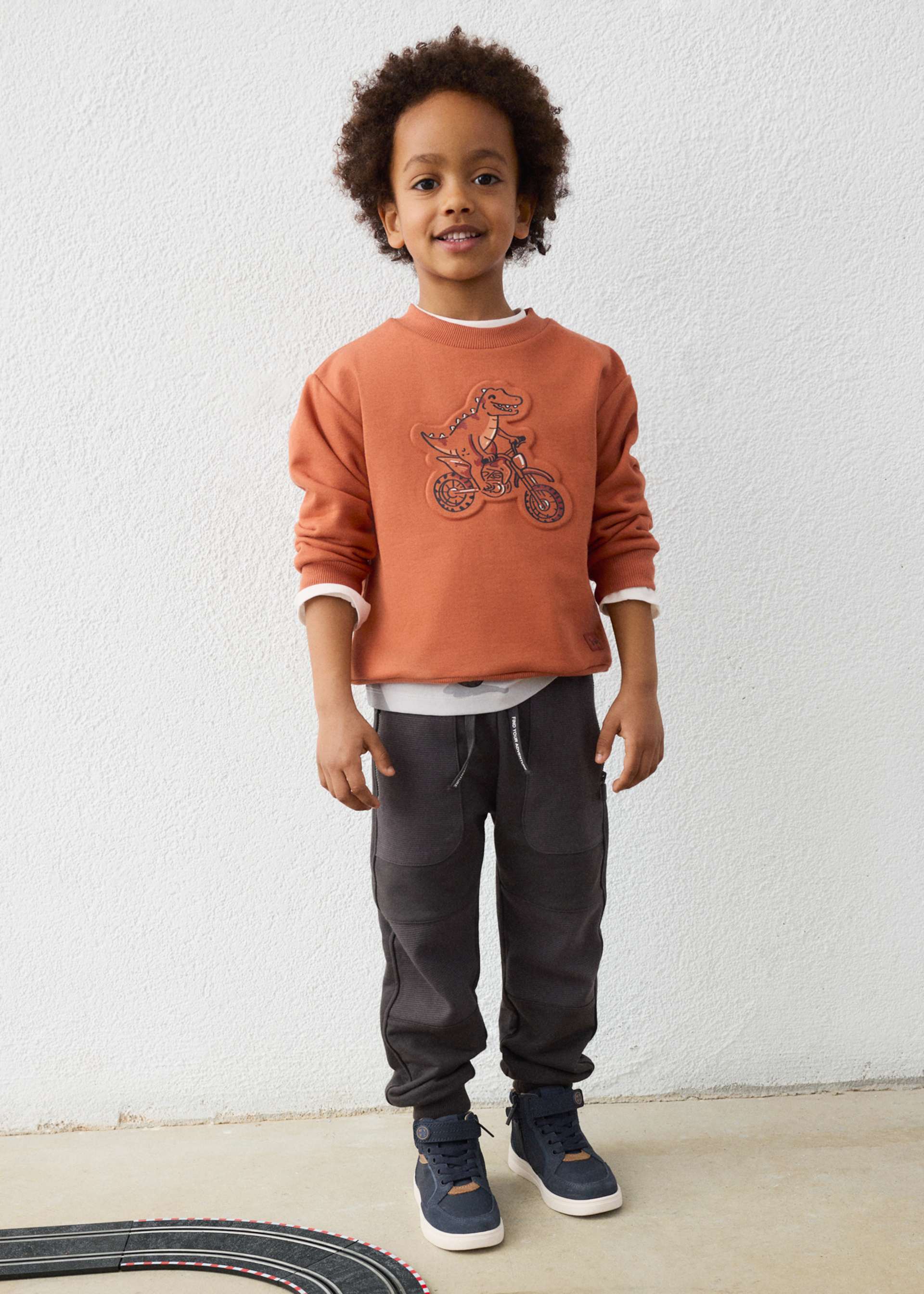 Child wearing a brown sweatshirt with a graphic design, black pants, and blue shoes against a white wall.