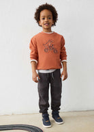 Child wearing a brown sweatshirt with a graphic design, black pants, and blue shoes against a white wall.