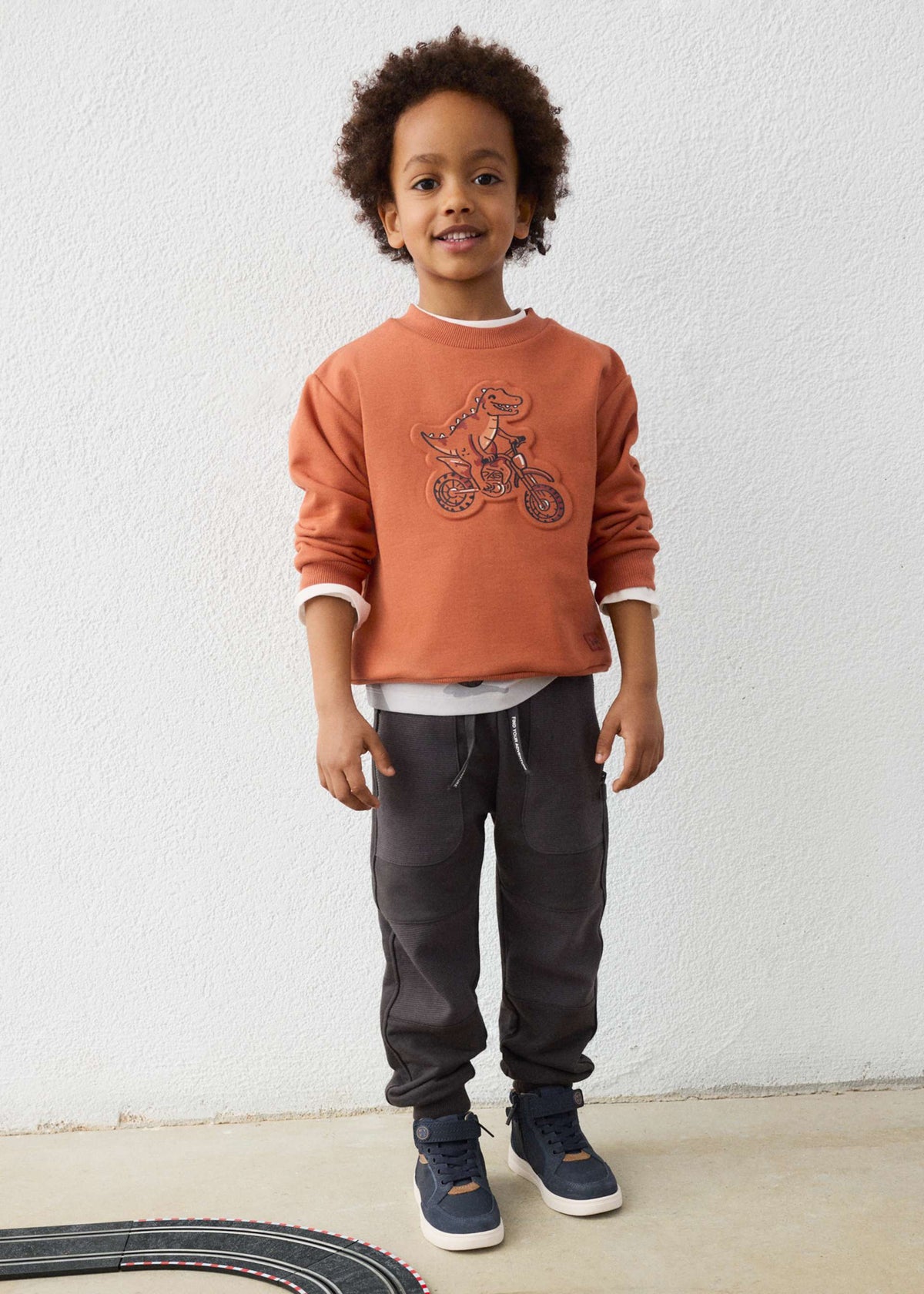 Child wearing a brown sweatshirt with a graphic design, black pants, and blue shoes against a white wall.