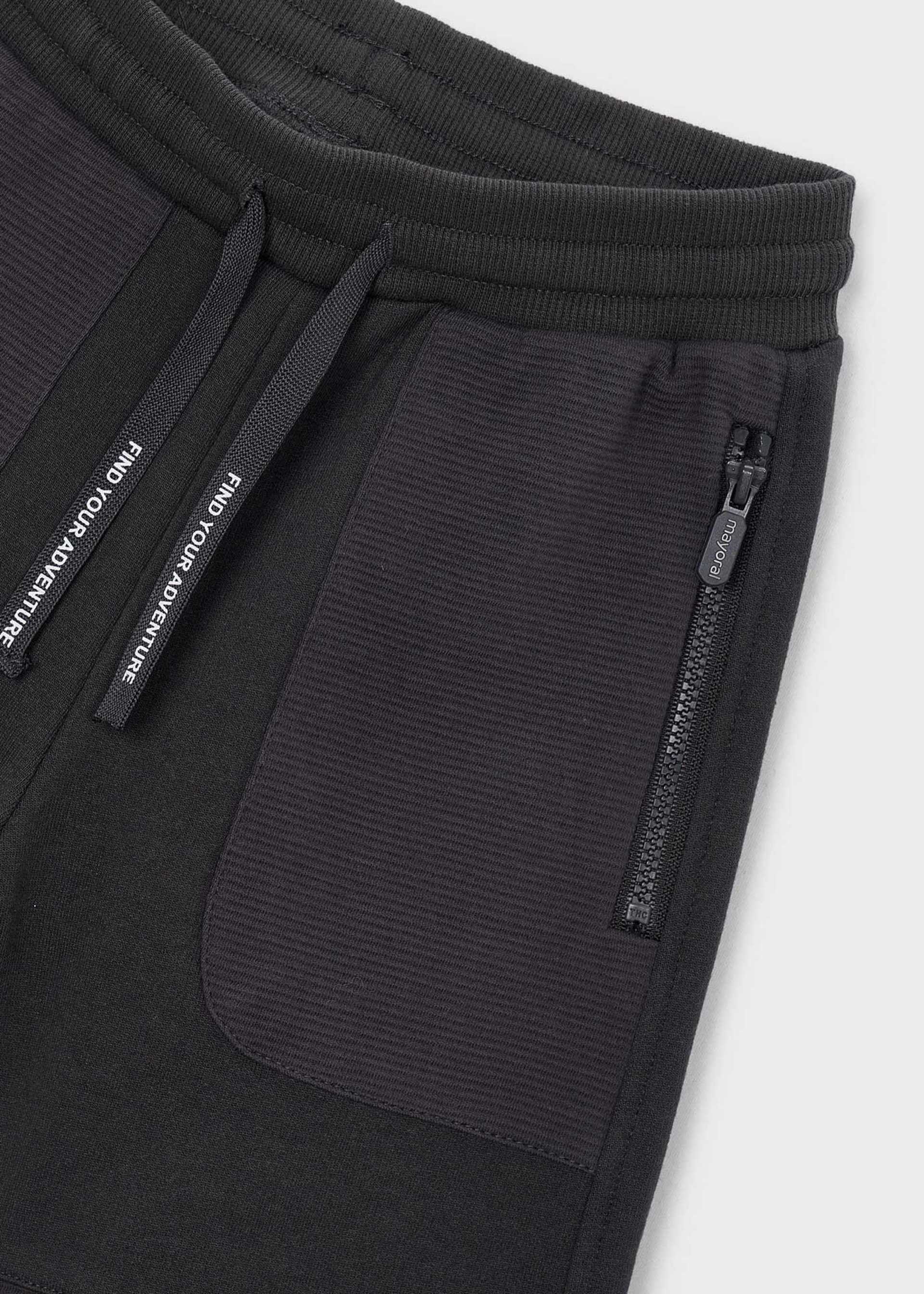 Close-up of black ribbed sweatpants showing a side pocket with a vertical zip, a drawstring with white text, and a ribbed waistband on a light gray background.
