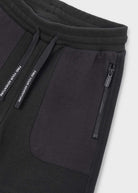 Close-up of black ribbed sweatpants showing a side pocket with a vertical zip, a drawstring with white text, and a ribbed waistband on a light gray background.