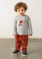 A toddler wearing a pair of brick-red Mayoral Baby Corduroy Knit Trousers paired with a grey graphic long-sleeve tee and casual sneakers.