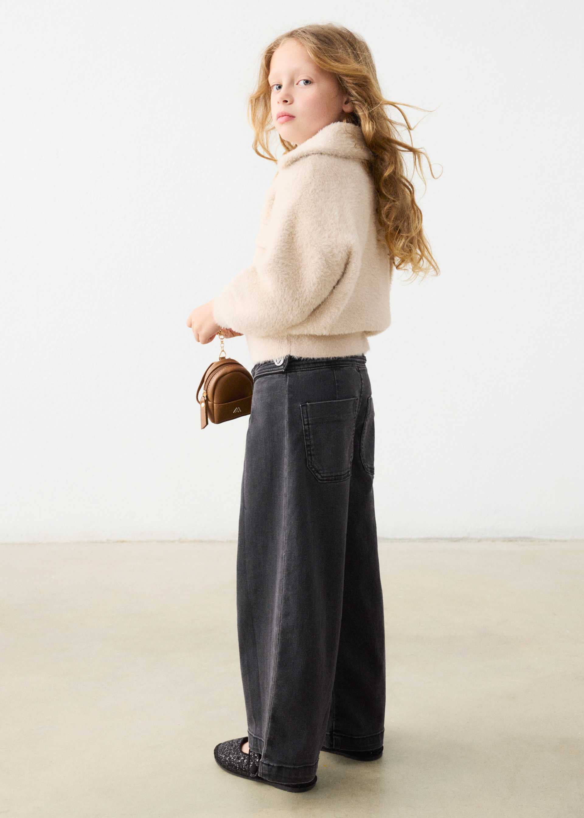 A young girl with long wavy light brown hair stands in profile wearing a cream fuzzy cropped sweater, dark charcoal wide-leg jeans, and black loafers, holding a small brown leather handbag against a plain white studio background.