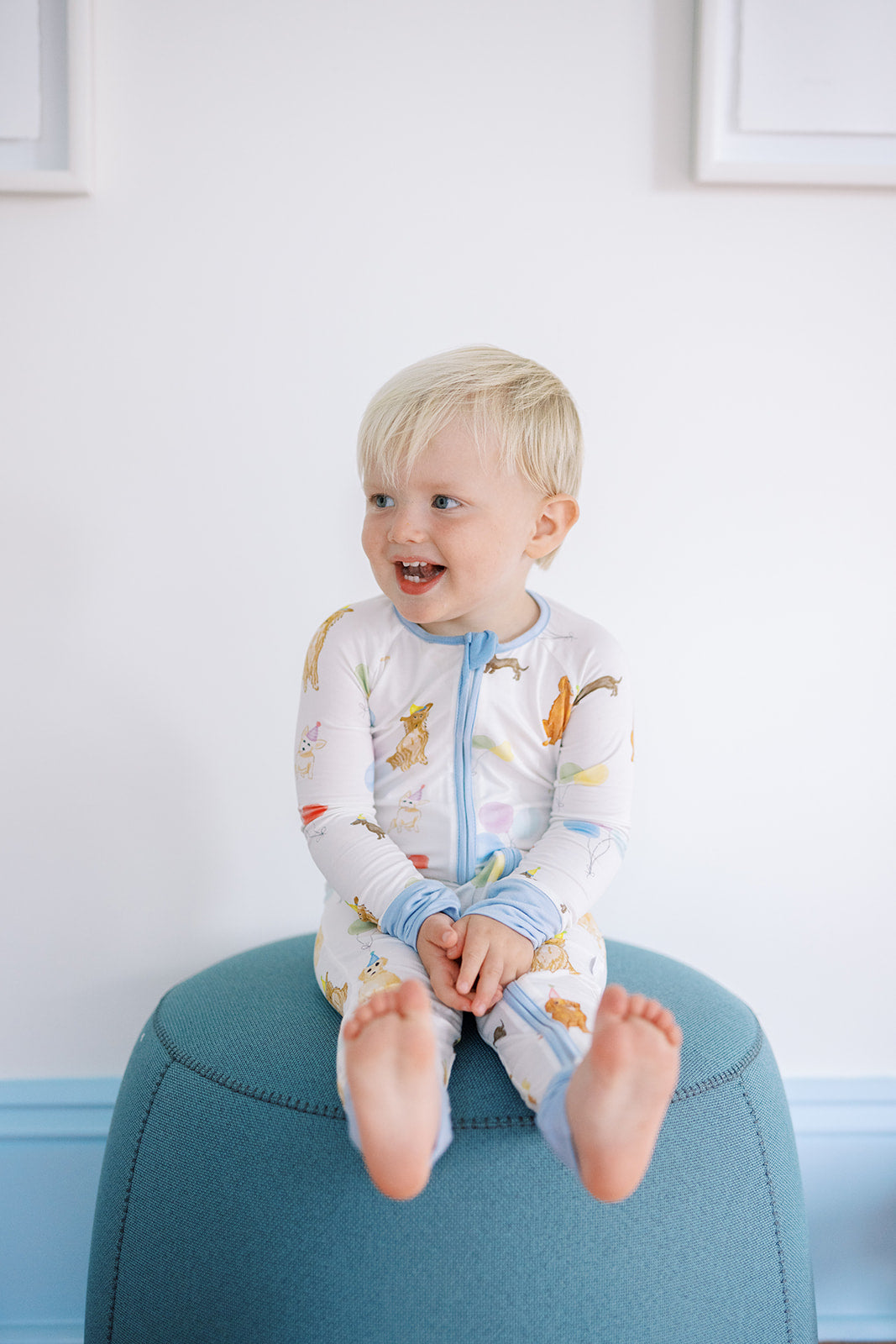Child wearing a colorful onesie sitting on a blue stool against a white wall.