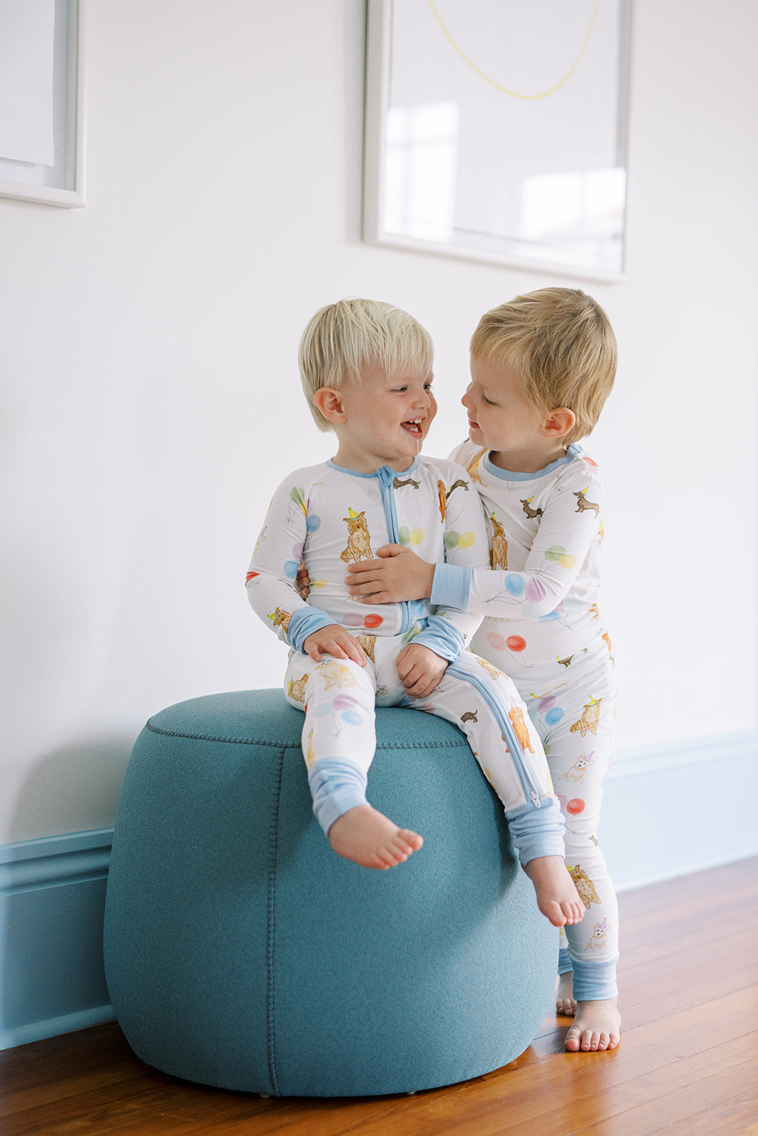 Two children in pajamas sitting on a blue ottoman in a room with white walls.