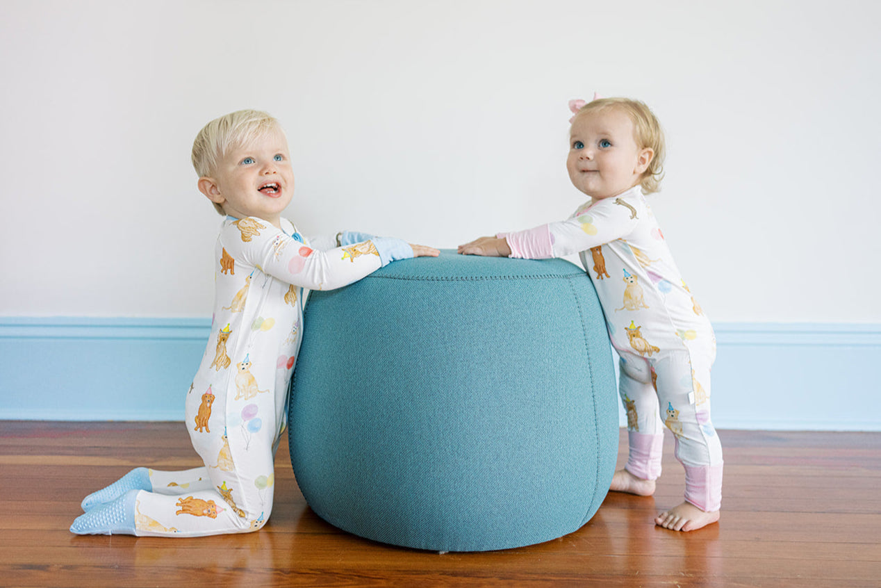 Two children playing with a blue ottoman in a room with a white wall and wooden floor.