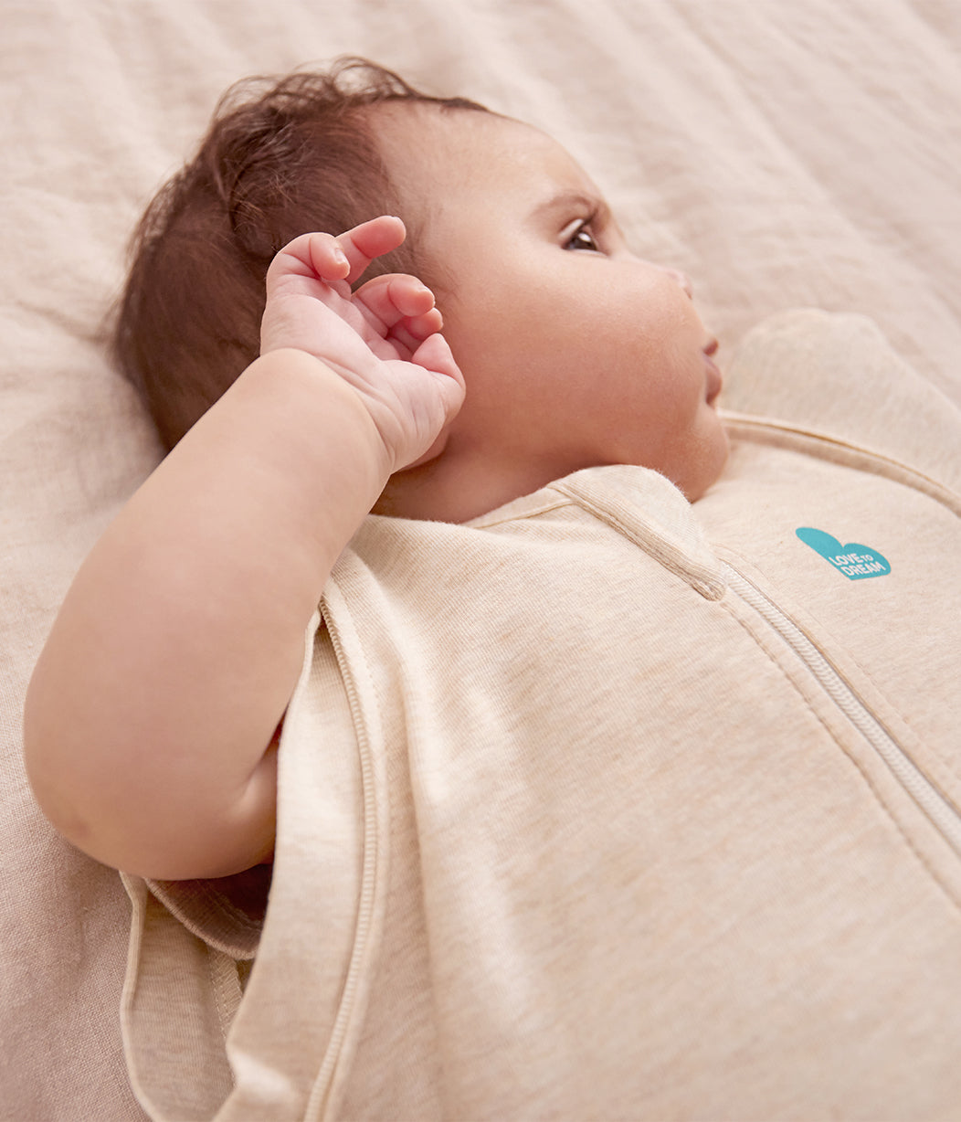 Baby wearing a beige swaddle with a visible brand logo on a soft, light-colored surface.