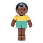 A wooden toy figure of a child with dark brown skin, black hair, a yellow top, and a mint green skirt, isolated on a white background.
