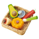 A set of wooden toy vegetables and fruit arranged in a small woven wicker basket on a plain white background. The set includes an avocado half, a red carrot, a green scallion, a yellow-orange pumpkin, a pear with a brown apple on top, and a small gray mushroom.