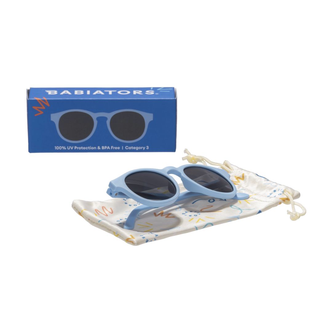 Children's sunglasses with blue frame and dark lenses, accompanied by a matching case and Babiators box on a white background.