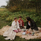 Two children and a woman sitting on a picnic blanket on the grass outdoors, enjoying a spread of food and drinks while surrounded by trees.
