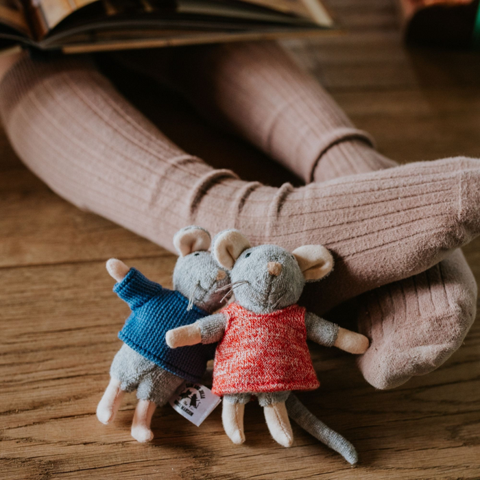 Two small mouse toys wearing sweaters on a wooden surface with a person's feet in socks in the background.