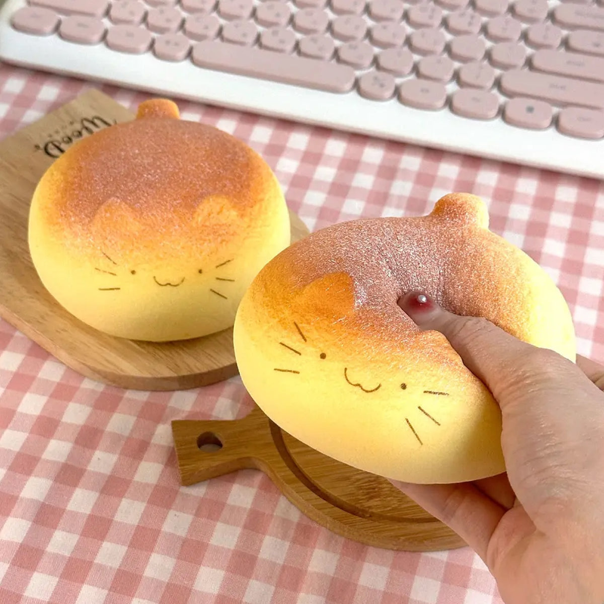 Two round, cat-face-shaped bread rolls on wooden serving boards, with one being pressed by a hand. The bread has a golden-brown baked top and a pale yellowish-beige bottom, with simple whisk and ear line drawings on the face.