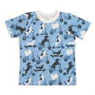 A short-sleeve kids' t-shirt with a light blue background featuring a repeating pattern of black and white dogs riding scooters. The shirt has a cream-colored crew neck collar.