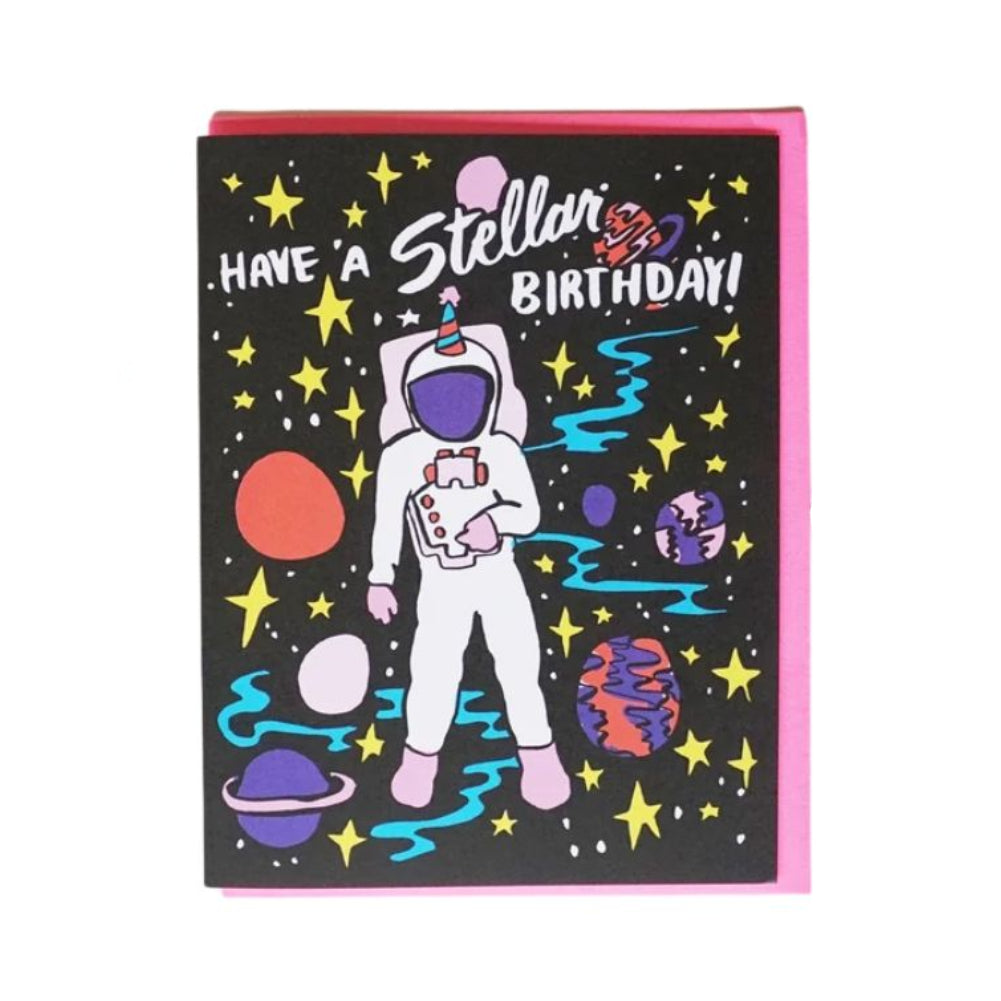 A square birthday card with a black space-themed design featuring an astronaut, planets, stars, and the text 'HAVE A Stellar BIRTHDAY!' in white and pink. The card has a pink border and is photographed on a white background