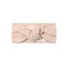 A pale blush pink fabric headband with a large twisted knot detail at the center, featuring a small white brand tag that reads 'MINDY MAC'. The product is photographed flat against a solid white background.