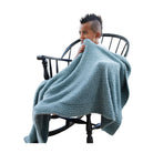 A child sitting in a black wooden rocking chair, wrapped in a plush, textured dusty blue throw blanket against a solid white background.