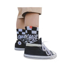 A close-up of a child's legs wearing black high-top sneakers with white laces and socks featuring a black and white checkerboard pattern and the word 'CHILL' in white script.