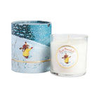 A white wax candle in a clear glass jar next to its cylindrical illustrated packaging. The packaging and label feature a winter scene with a bear in a yellow coat walking through snow, and the label reads 'First Snowfall in the Pines'.