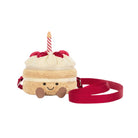 A plush cake-shaped crossbody bag with cream-colored frosting, red cherries, a red ribbon bow, a red strap, and a small red-and-white striped candle on top, shown on a plain white background.