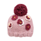 A pink knit beanie hat with a large dark red pom-pom on top and crocheted flower appliqués in dark red, white, and yellow.