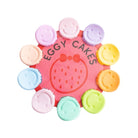 A circular pink display card reading 'EGGY CAKES' with a strawberry illustration, surrounded by eight small pastel round items with smiley faces in pink, purple, peach, blue, mint, green, orange, and yellow, all on a white background.