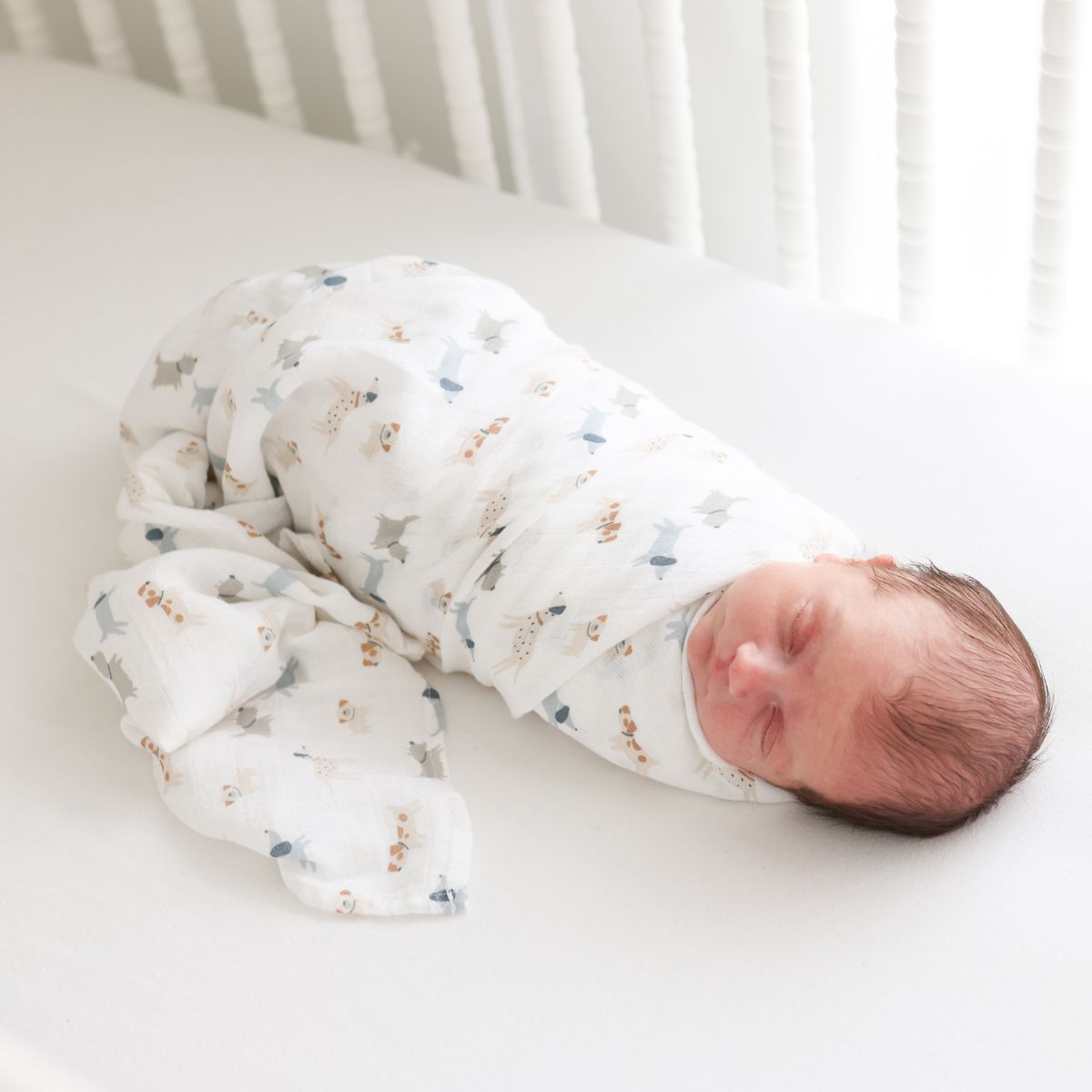 Newborn baby swaddled in a white blanket with small patterns, lying on a white surface.