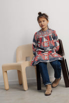A young child sitting on a black chair next to a light-colored chair in a minimalist room, wearing The Quill Dress by The New Society with dark pants and boots.